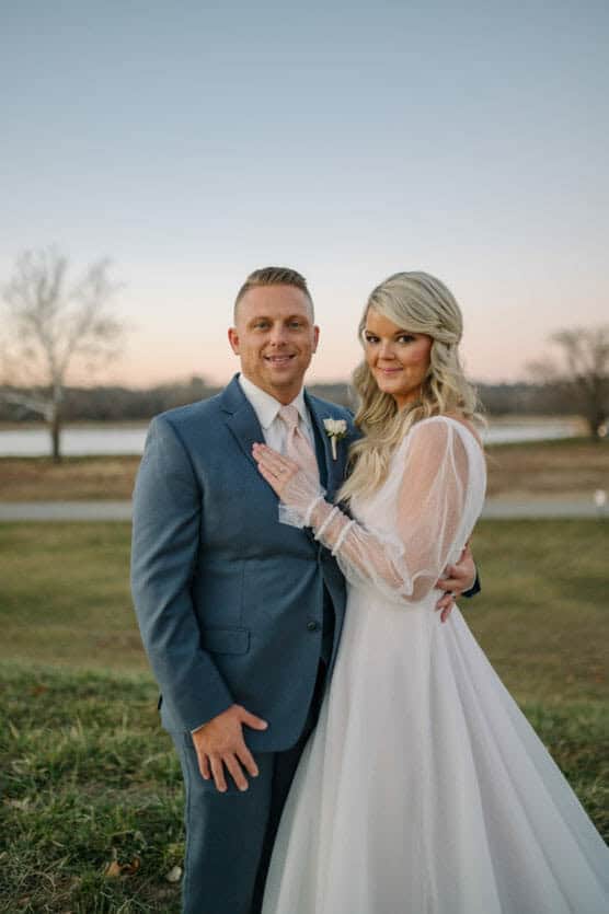 A bride and groom stand outdoors on grass at sunset, smiling at the camera. The bride wears a long white dress and the groom wears a blue suit. Trees and water are visible in the background. by Justin Salem Meyer