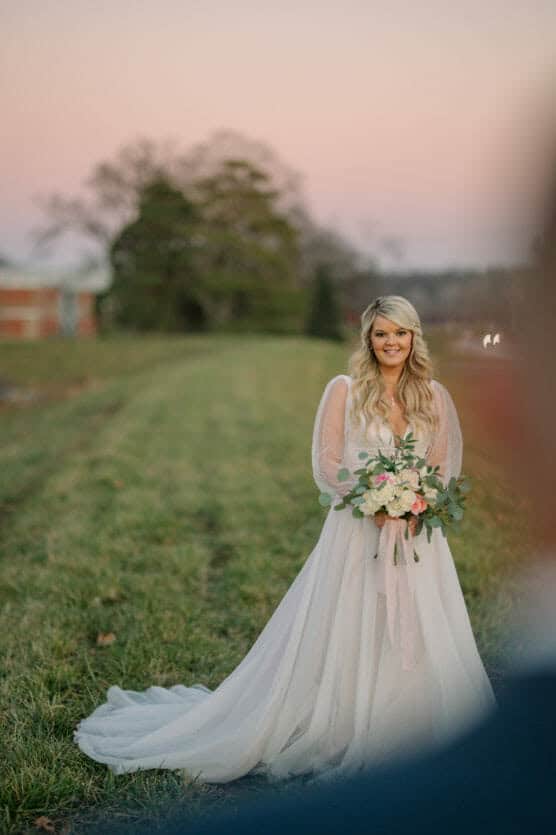 A bride in a white gown holds a bouquet of flowers and smiles while standing on a grassy field at sunset, with soft pink and purple tones in the sky and blurred trees in the background. by Justin Salem Meyer