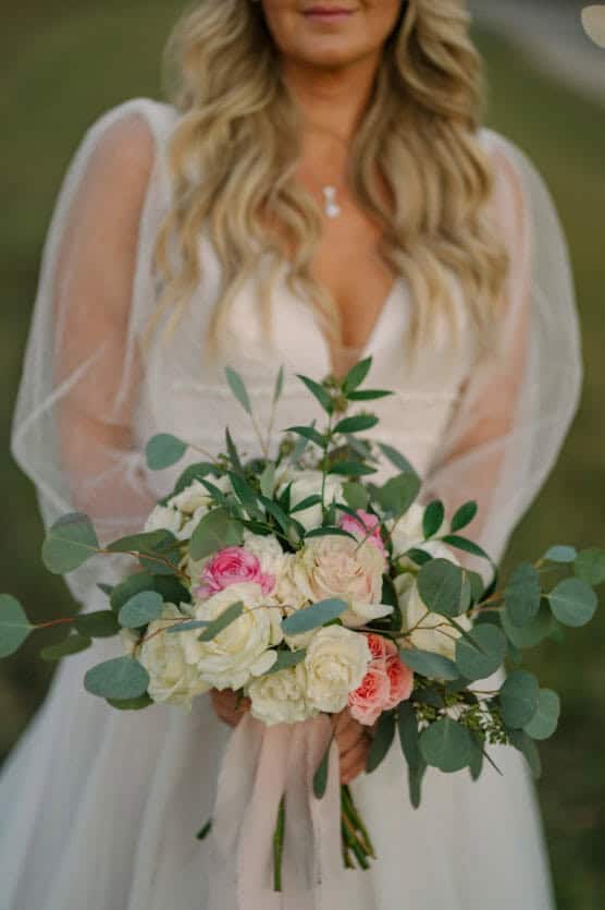 A bride in a white dress with long, sheer sleeves holds a bouquet of white, pink, and peach roses mixed with greenery. Her long, wavy blonde hair hangs down, and she wears a delicate necklace. by Justin Salem Meyer