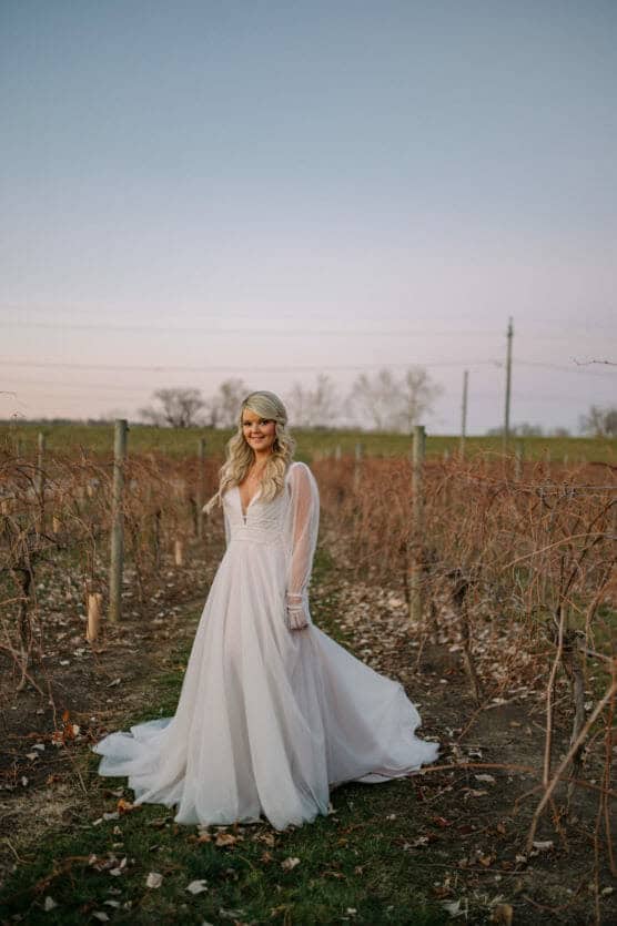 A woman in a long white wedding dress stands in a vineyard at sunset, surrounded by bare grapevines and fallen leaves, with a soft blue and pink sky in the background. by Justin Salem Meyer