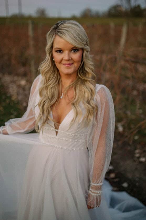 A woman with long, wavy blonde hair wearing a white, sheer-sleeved wedding dress stands outdoors, holding her dress and smiling, with a blurred natural background. by Justin Salem Meyer