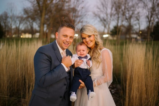 A smiling man and woman, dressed formally, stand outdoors holding a baby in a bow tie. The baby looks upset. Tall grass and bare trees are in the background, suggesting early spring or late autumn. by Justin Salem Meyer