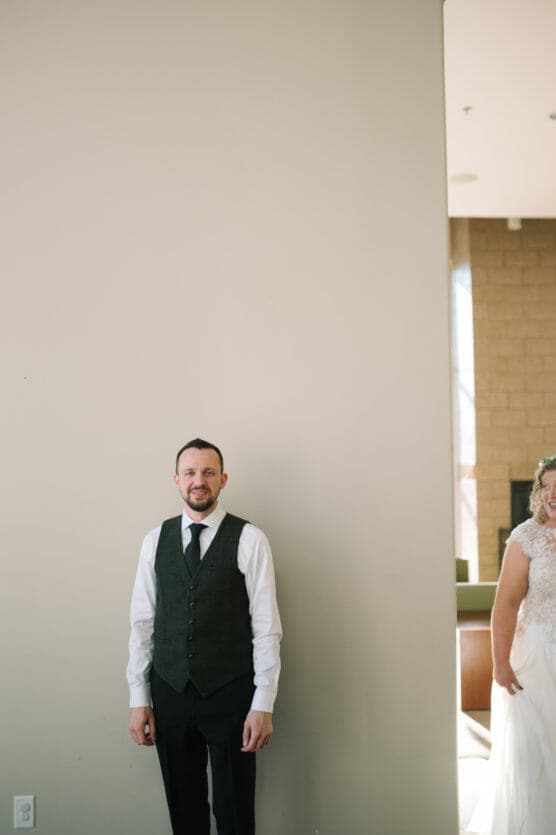 A man in a vest and tie stands on the left side of a beige wall, smiling. On the right, a woman in a white wedding dress is partially visible, mostly hidden behind the wall, in a well-lit room. by Justin Salem Meyer