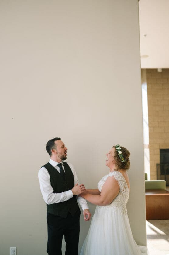A bride and groom stand indoors, facing each other and laughing joyfully. The bride wears a white dress with lace details and a flower crown, while the groom wears a black vest and tie over a white shirt. by Justin Salem Meyer