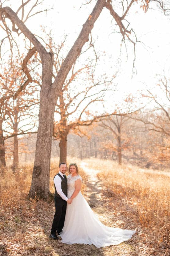 A bride and groom stand hand in hand on a sunlit woodland path, surrounded by tall trees with sparse autumn leaves and golden grass, smiling at the camera. by Justin Salem Meyer
