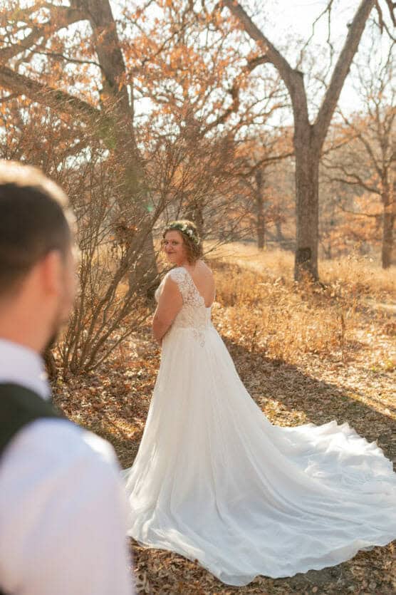 A bride in a white lace wedding dress stands outdoors in autumn, looking over her shoulder at a groom in the foreground. Sunlight filters through bare trees and fallen leaves cover the ground. by Justin Salem Meyer
