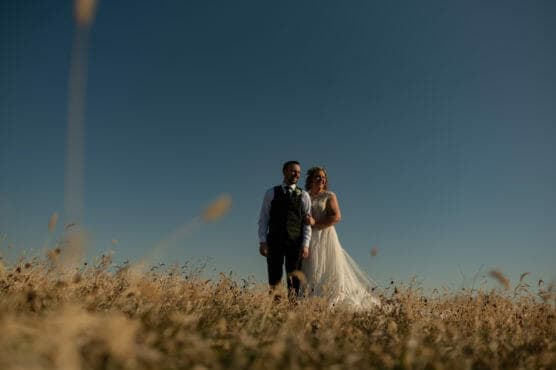 A bride and groom stand together in a field of tall, golden grass under a clear blue sky, smiling and holding hands on their wedding day. by Justin Salem Meyer