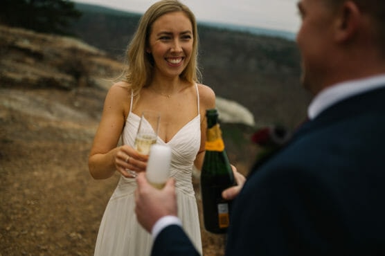 A woman in a white dress smiles while holding a champagne glass, as a man in a suit offers to pour champagne outdoors with a scenic, rocky landscape in the background. by Justin Salem Meyer