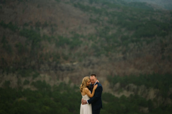 A couple embraces and smiles in formal attire on a green mountainside, with blurred forested hills in the background under overcast skies. by Justin Salem Meyer