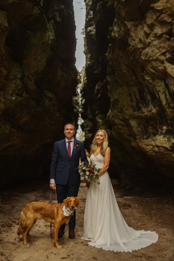 A bride in a white dress and a groom in a navy suit stand together holding a bouquet, with a golden retriever on a leash, set between two tall, rocky cliffs. by Justin Salem Meyer