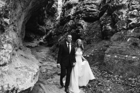 A bride and groom walk hand in hand along a rocky path outdoors, surrounded by large rock formations. The bride wears a flowing white dress and the groom wears a black suit. The scene is in black and white. by Justin Salem Meyer