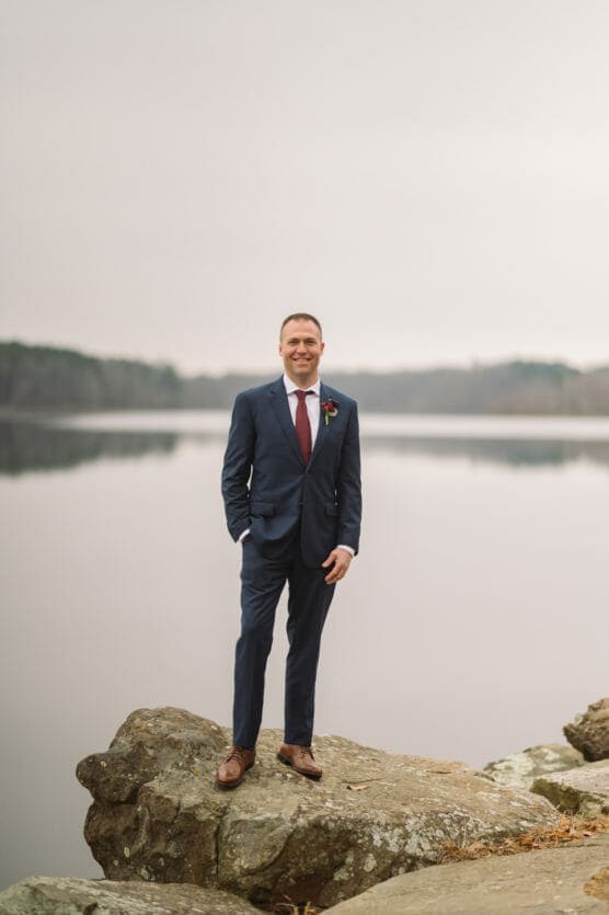 A man in a blue suit with a red tie stands smiling on large rocks beside a calm lake. The background shows still water and distant trees under an overcast sky. by Justin Salem Meyer