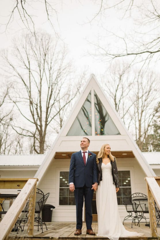 A bride and groom stand holding hands on the porch of an A-frame house. The groom wears a navy suit and red tie, while the bride wears a white dress and a black jacket. Bare trees and patio furniture surround them. by Justin Salem Meyer