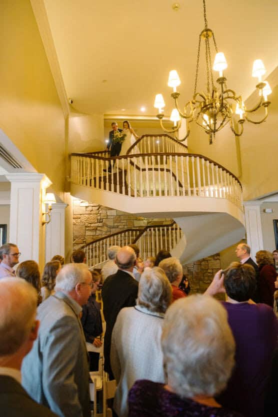A bride and groom stand at the top of a spiral staircase, smiling at guests gathered below in a warmly lit room with a large chandelier. The guests look up in anticipation. by Justin Salem Meyer
