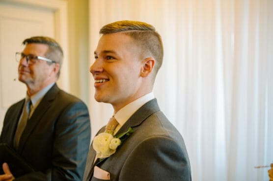 A young man in a gray suit with a white rose boutonniere smiles while standing indoors next to an older man in a suit, with light streaming through sheer curtains in the background. by Justin Salem Meyer