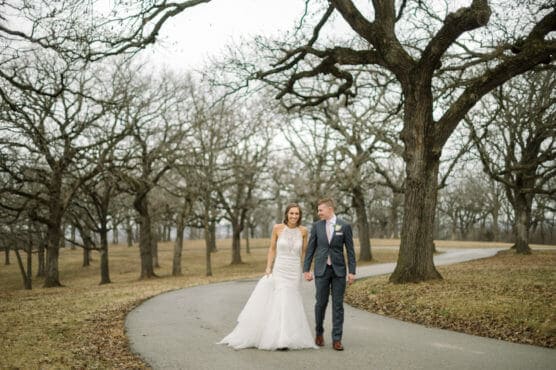 A bride in a white wedding dress and a groom in a suit walk hand-in-hand on a winding path through a grove of leafless trees on a cloudy day. by Justin Salem Meyer