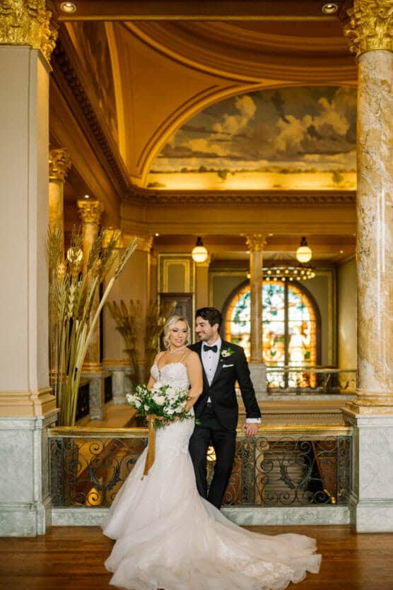 A bride in a white gown and a groom in a black tuxedo stand together in an ornate hall with marble columns, golden decor, and a large stained glass window in the background. by Justin Salem Meyer