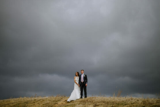 A bride and groom stand close together on a grassy hill under a dramatic, overcast sky. The landscape is open and minimal, highlighting the couple against the dark, cloudy backdrop. by Justin Salem Meyer