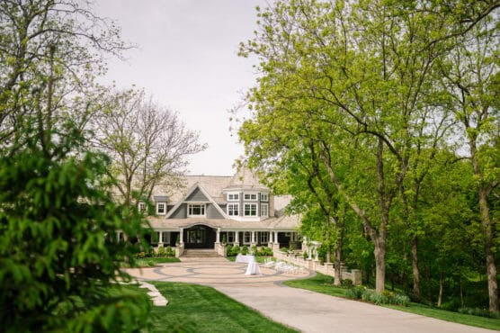 A large house with a grey roof and white trim sits at the end of a curved driveway, surrounded by lush green trees and manicured lawns on a bright day. A circular stone patio and two small cocktail tables are near the entrance. by Justin Salem Meyer