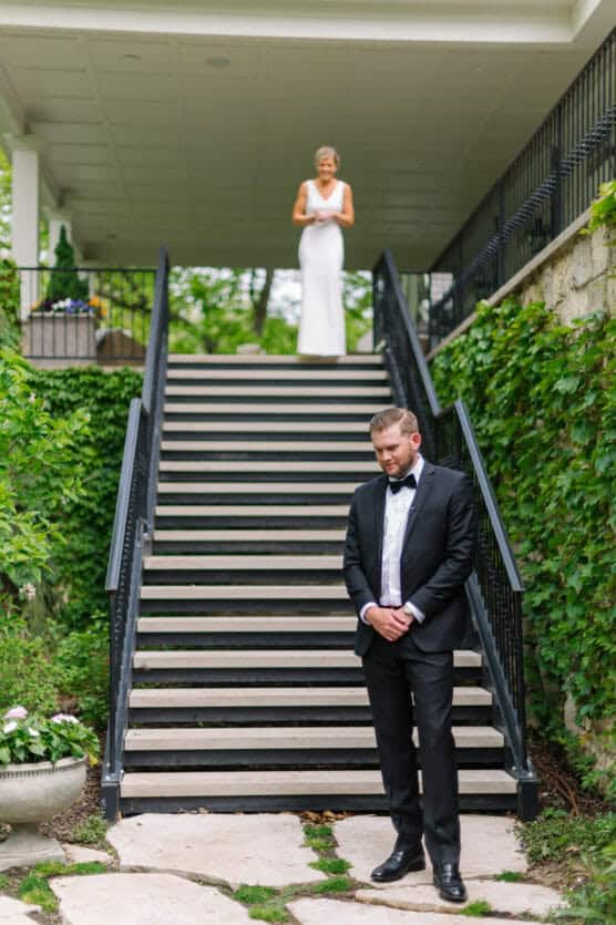 A groom in a black suit stands with his head bowed at the bottom of outdoor stairs, while a bride in a white dress stands at the top, both surrounded by greenery. by Justin Salem Meyer