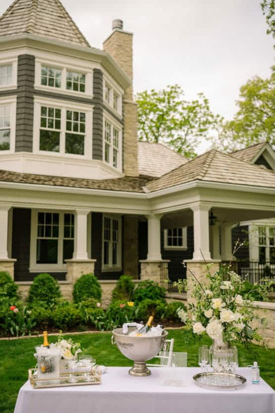 A table with champagne, glasses, and white floral arrangements set up outdoors in front of a large, elegant house with gray siding, white trim, and stone accents. Lush greenery surrounds the scene. by Justin Salem Meyer