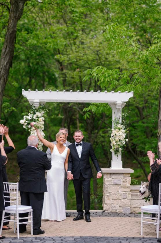A bride in a white dress and a groom in a black tuxedo smile and hold hands under a floral arch as wedding guests applaud outdoors amid green trees. by Justin Salem Meyer