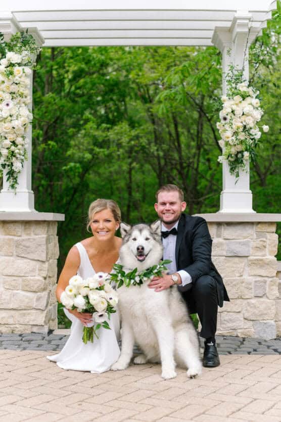 A bride and groom kneel and smile beside a fluffy white dog wearing a green garland collar. They are outdoors under a decorated white gazebo with white flowers, with green trees in the background. by Justin Salem Meyer