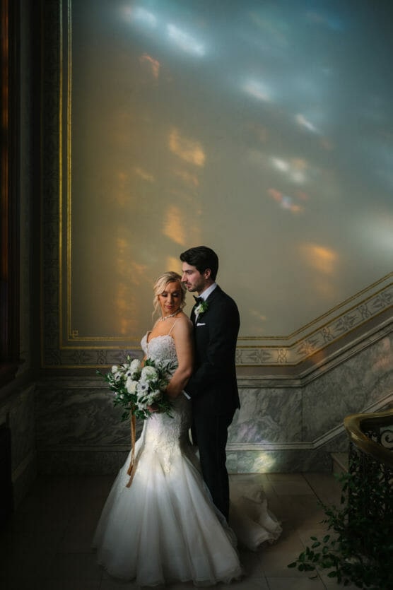 A bride and groom stand closely together on a marble staircase, bathed in soft, colorful light reflections, with the bride holding a white bouquet and both dressed in formal wedding attire. by Justin Salem Meyer
