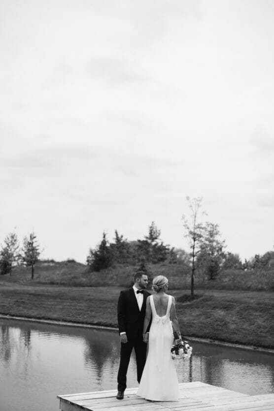 A bride and groom stand on a dock by a calm pond, facing each other. The bride holds a bouquet and wears a backless dress. The groom wears a tuxedo. Trees and grass line the background under a cloudy sky. Black and white photo. by Justin Salem Meyer
