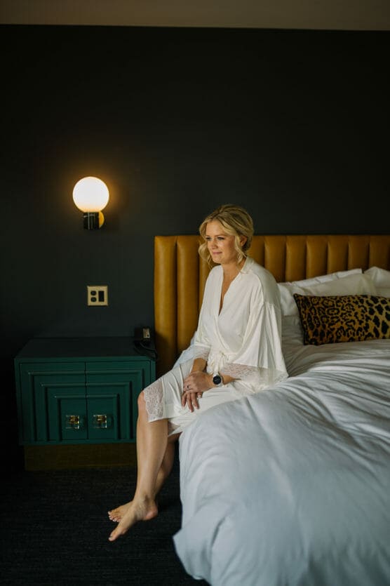 A woman in a white robe sits on the edge of a bed with white linens, next to a teal nightstand and a decorative pillow with a leopard print cover, against a dark wall with a round lit wall sconce. by Justin Salem Meyer