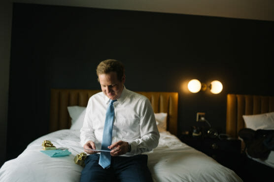 A man in a white dress shirt and blue tie sits on a bed, looking down and smiling at a card in his hands. There are cards and a small gift beside him. The room has dark walls and modern lighting. by Justin Salem Meyer