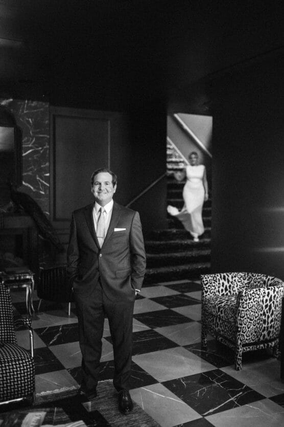 A man in a suit stands smiling in a stylish, black-and-white room with checkered floors; in the background, a woman in a white dress descends a staircase, slightly out of focus. by Justin Salem Meyer