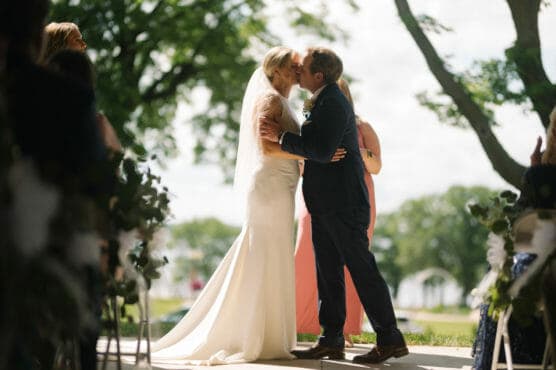 A bride and groom share a kiss at their outdoor wedding ceremony, dressed in a white gown and dark suit, surrounded by guests and greenery on a sunny day. by Justin Salem Meyer