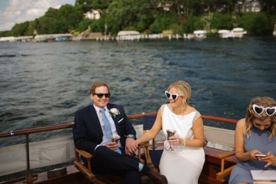 A couple in formal attire with heart-shaped sunglasses sit on a boat, smiling and holding drinks, while another woman next to them looks at her phone. A lake and shoreline with houses and trees are in the background. by Justin Salem Meyer