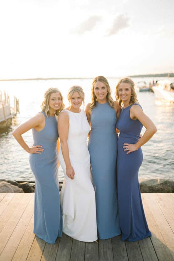 Four women stand together on a wooden dock by the water at sunset. The woman second from left wears a white dress, while the others wear matching blue dresses, smiling with their arms around each other. by Justin Salem Meyer
