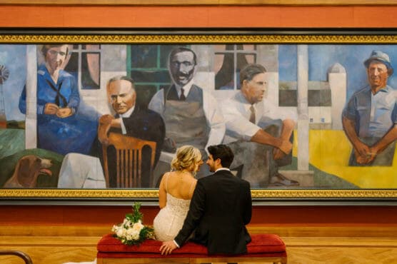 A bride and groom sit closely on a red bench, facing a large, colorful mural of five men and a dog, in an art gallery. The bride holds a bouquet as they look at the artwork together. by Justin Salem Meyer