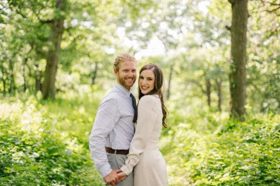 A couple stands close together in a sunlit, green forest, smiling at the camera while holding hands. The woman wears a light dress and the man wears a shirt and tie. The background is lush with trees and foliage. by Justin Salem Meyer