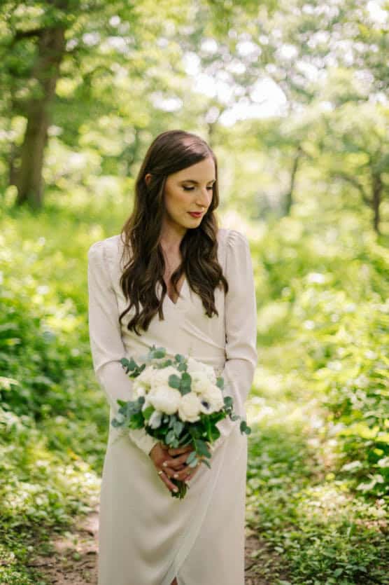 A woman in a long-sleeved ivory dress stands outdoors in a sunlit, green forest, holding a bouquet of white flowers and greenery while looking down thoughtfully. by Justin Salem Meyer