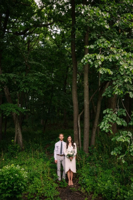 A couple stands together in a lush, green forest. The man wears a light shirt and tie, and the woman wears a light dress and holds a bouquet. Tall trees and dense foliage surround them. by Justin Salem Meyer