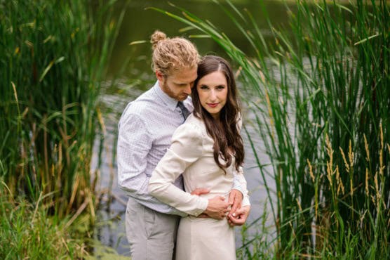 A couple stands close together by a pond, surrounded by tall green reeds. The man, in a light shirt and gray pants, embraces the woman from behind. She wears a cream dress and looks at the camera, while he looks down lovingly. by Justin Salem Meyer