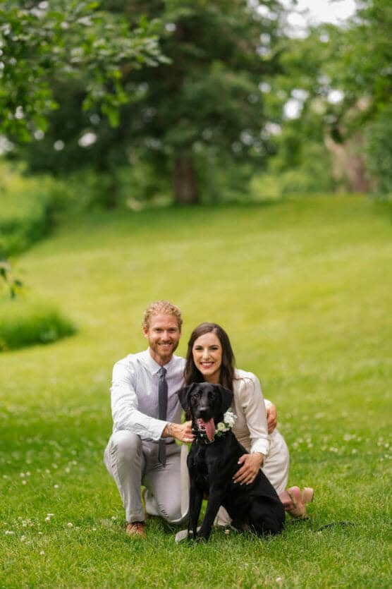 A smiling couple dressed in light-colored formal clothes kneels on grass, posing with a black dog wearing a flower collar, in a lush green outdoor setting. by Justin Salem Meyer