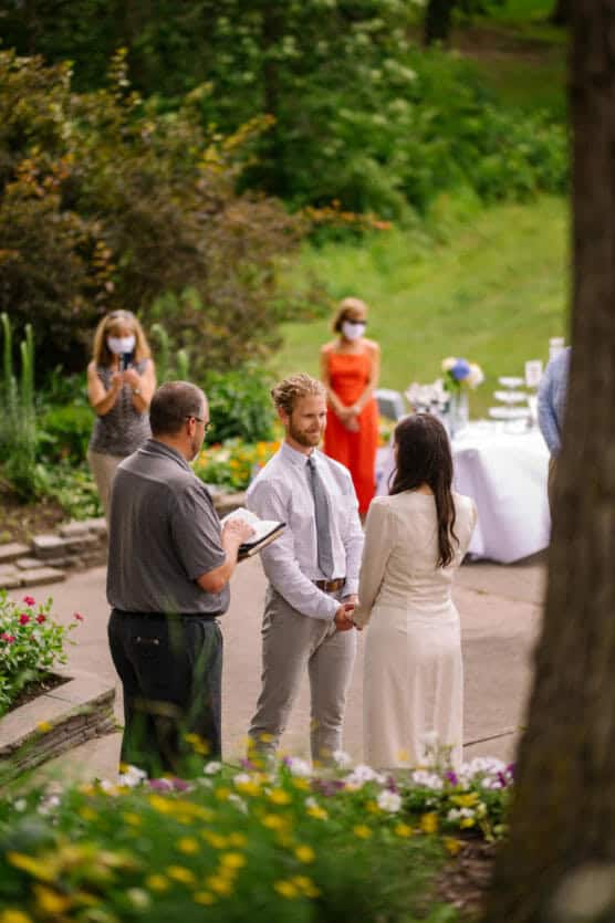 A couple stands facing each other holding hands during an outdoor wedding ceremony, with a person officiating. Guests in the background wear face masks and stand apart, surrounded by greenery and flowers. by Justin Salem Meyer