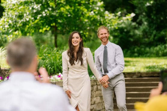 A smiling couple stands outdoors holding hands, dressed in semi-formal clothes, with greenery and stone steps in the background and people taking photos of them in the foreground. by Justin Salem Meyer