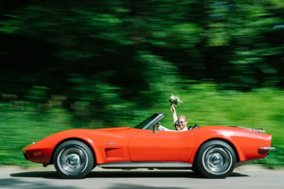 A joyful couple drives a classic red convertible sports car with the top down; the passenger raises a bouquet of flowers while greenery blurs in the background, suggesting motion and speed. by Justin Salem Meyer