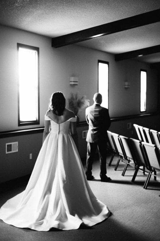 A bride in a flowing gown stands behind a man in a suit inside a sunlit room with empty chairs, captured in black and white. by Justin Salem Meyer