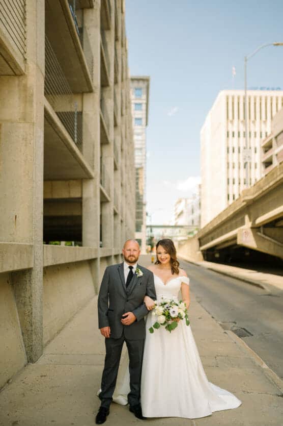 A bride in a white dress holding a bouquet stands beside a groom in a gray suit and black tie on a city sidewalk, with tall concrete buildings and a blue sky in the background. by Justin Salem Meyer