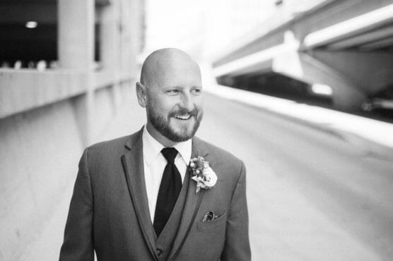 A man in a suit with a boutonniere on his lapel smiles while standing outdoors in an urban setting. The image is in black and white, and the background shows blurred architecture and a road. by Justin Salem Meyer