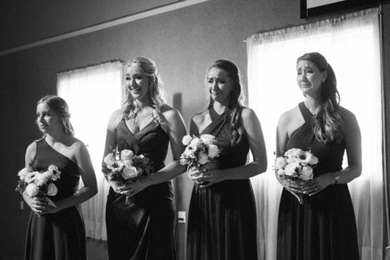 Four bridesmaids in matching dark dresses hold flower bouquets and stand side by side indoors, their expressions emotional as they watch something off camera. Sunlight streams through two windows behind them. by Justin Salem Meyer