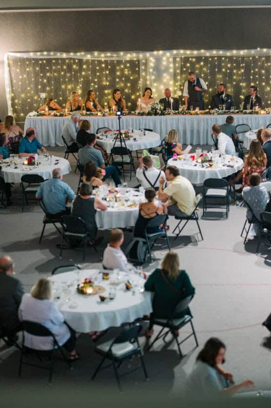 Guests are seated at round tables, watching a wedding reception head table where speeches are being given. String lights create a warm, festive backdrop behind the wedding party. by Justin Salem Meyer