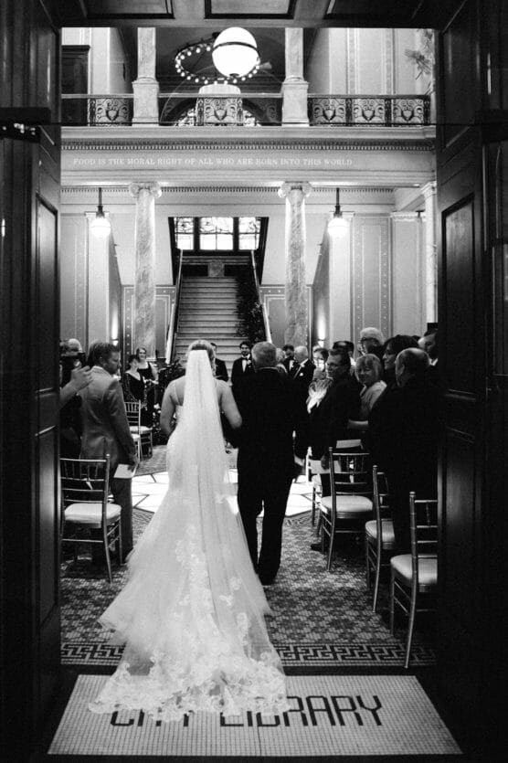 A bride in a long veil and gown walks down the aisle with an older man in a grand, ornate hall filled with seated guests, heading toward a staircase for a wedding ceremony. by Justin Salem Meyer